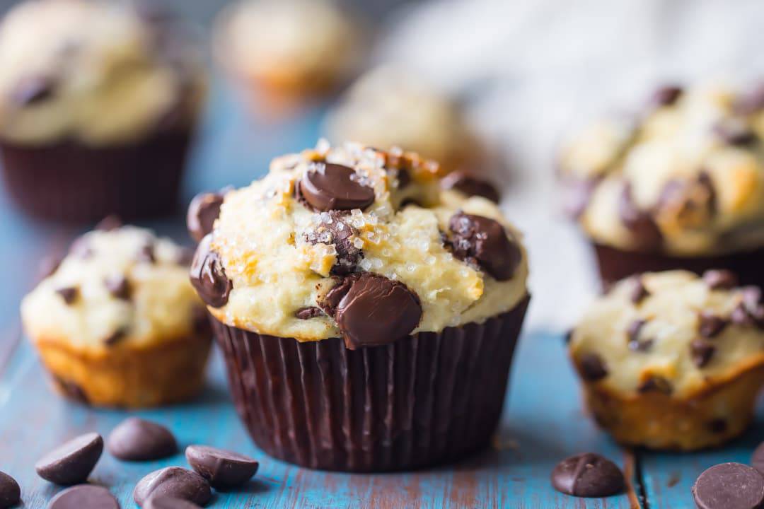 Horizontal image of a freshly baked chocolate chip muffin on an aqua-painted wood background, with chocolate chip mini-muffins in the background. 