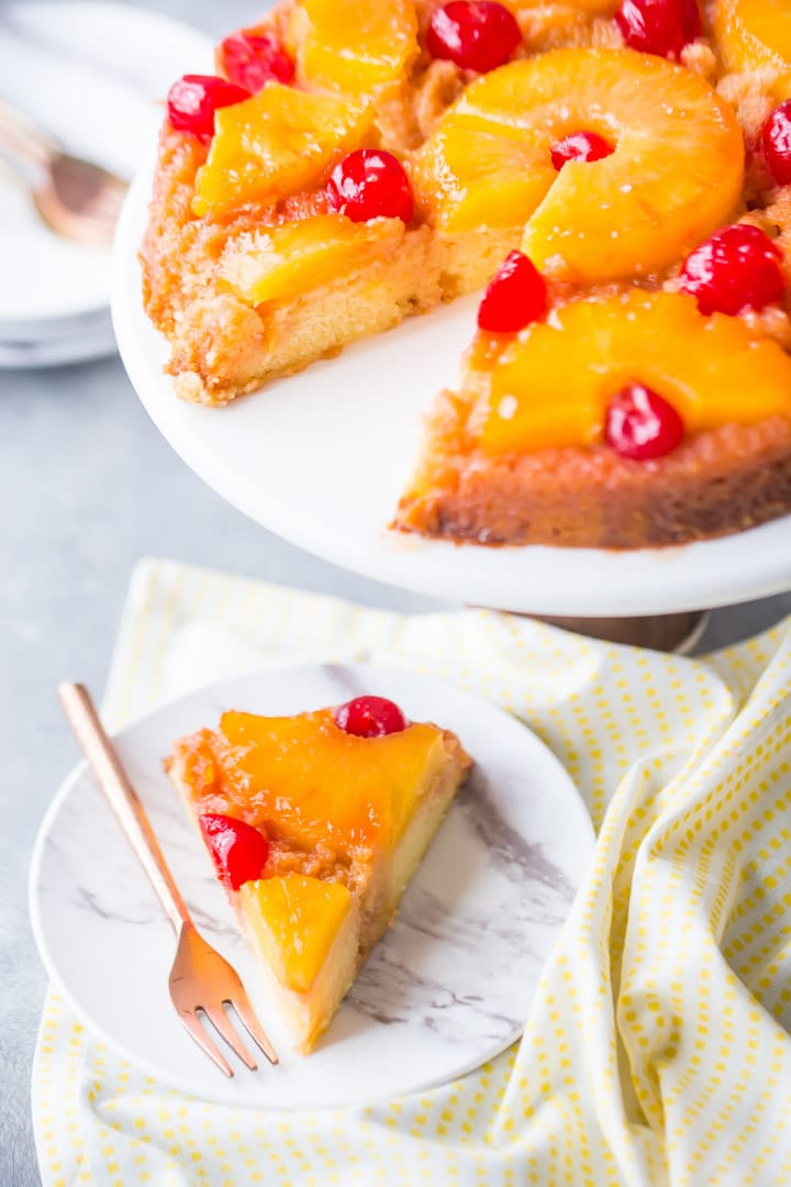 Vertical image of a pineapple upside down cake on a stand with a slice cut out and plated.