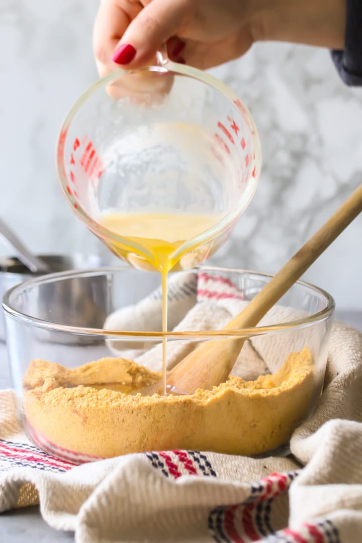 Vertical image of melted butter being poured into a bowl of graham cracker crumbs to make graham cracker crust.