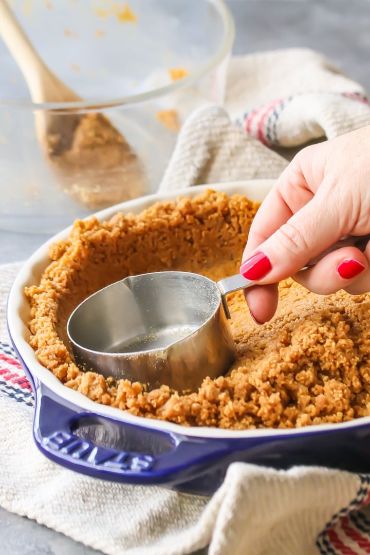 Vertical image of graham cracker crust being pressed into a pie dish with the bottom of a measuring cup.