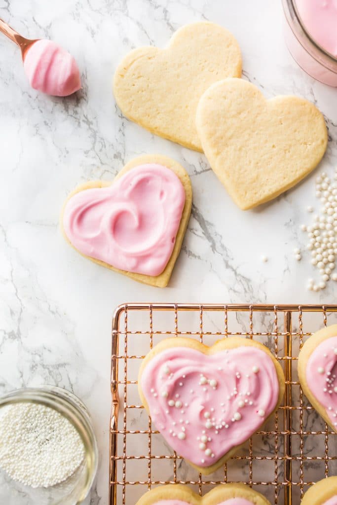Vertical image of heart-shaped cream cheese cut-out sugar cookies on a copper cooling rack with pink sugar cookie frosting.