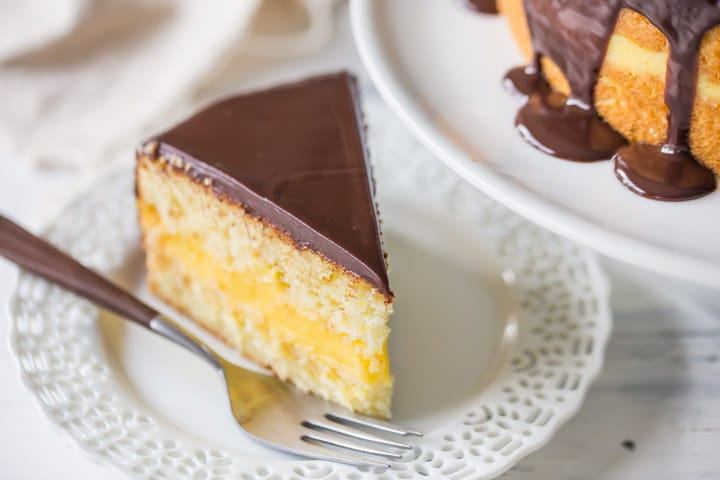 Perfect slice of Boston Cream Pie on a plate next to the full cake, showing ganache topping dripping down the sides.
