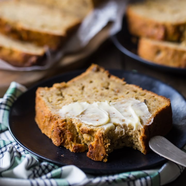 Square image of a slice of buttered zucchini bread on a dark plate.
