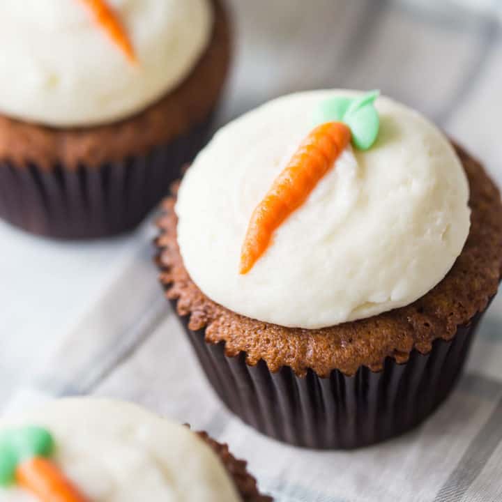 A carrot cake cupcake on a plaid cloth, decorated with cream cheese frosting and an orange carrot piped on top with a green leaf.