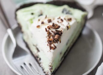 A slice of grasshopper pie with whipped cream and chocolate shavings, on a white plate with a green plaid napkin in the background.