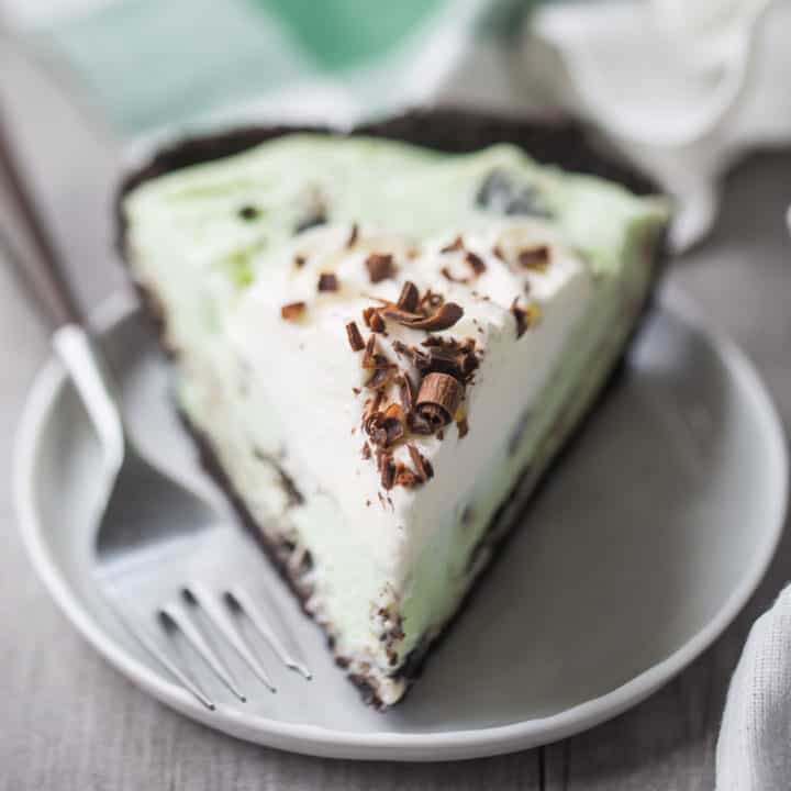 A slice of grasshopper pie with whipped cream and chocolate shavings, on a white plate with a green plaid napkin in the background.