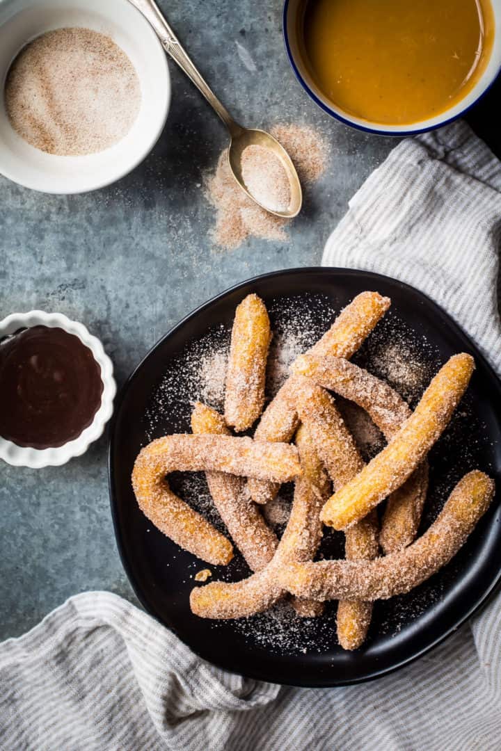Authentic Mexican churros on a dark plate, surrounded by bowls of dulce de leche, cinnamon sugar, and chocolate sauce.