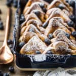 Baked french toast with powdered sugar in a blue casserole dish with a copper serving spoon