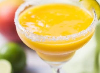 Frozen mango margarita in a salt-rimmed glass with mangoes and limes in the background.