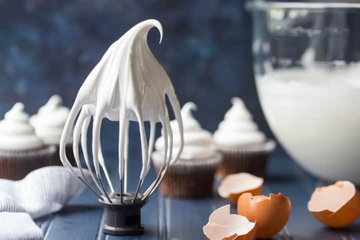 Whisk attachment with marshmallow frosting with egg shells and cupcakes in the background.