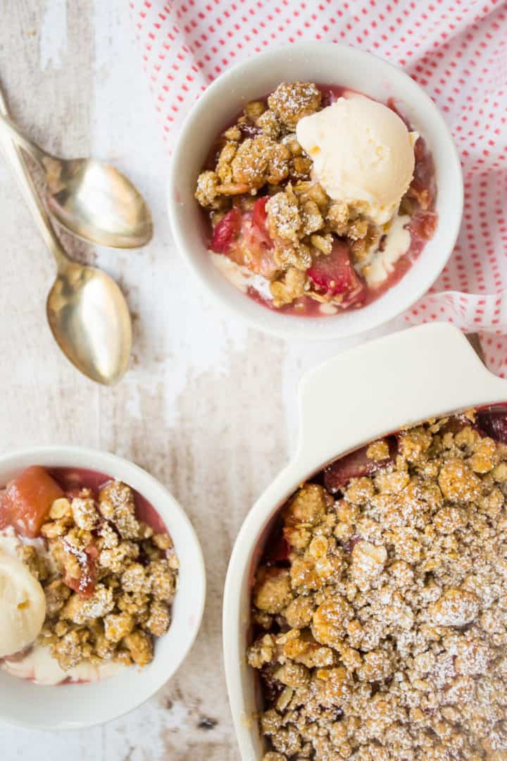 Overhead image of rhubarb crisp in a white ceramic baking dish, with two small bowls of rhubarb crisp and silver spoons.