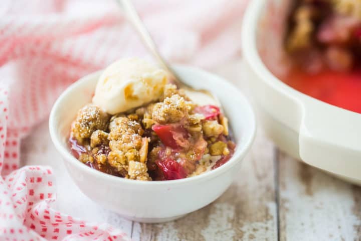 Bowl of homemade rhubarb crisp with vanilla ice cream and a baking dish in the background.