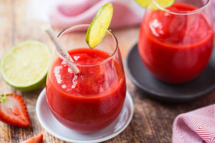 Horizontal image of 2 frozen strawberry daiquiris with cut limes and strawberries in the background.