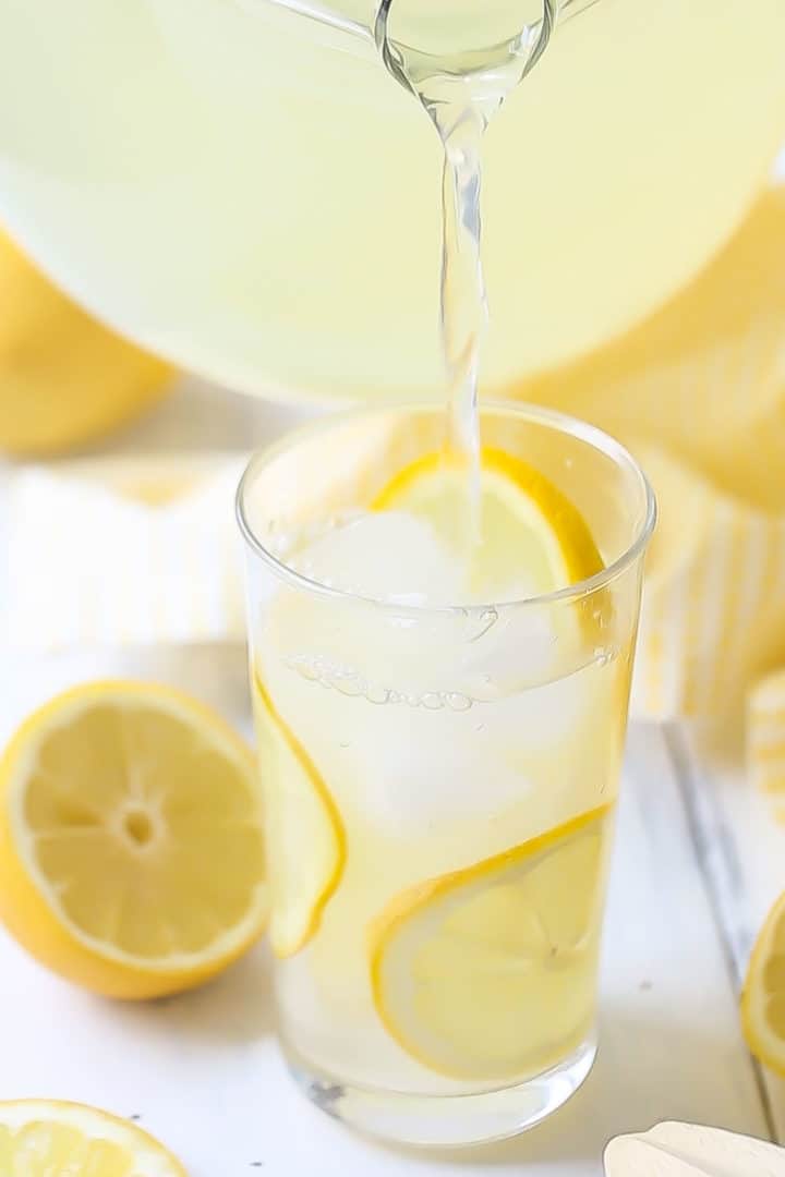Pouring lemonade from a glass pitcher into a tall glass of ice with lemon slices.