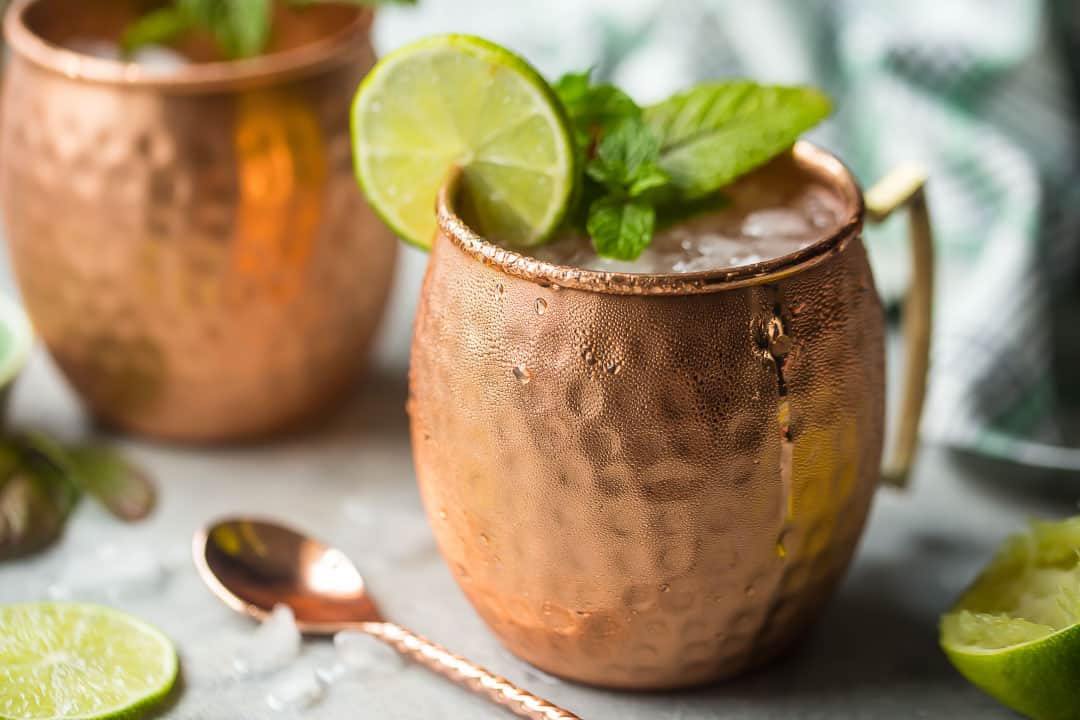Two copper Moscow mule mugs surrounded by crushed ice and fresh limes.