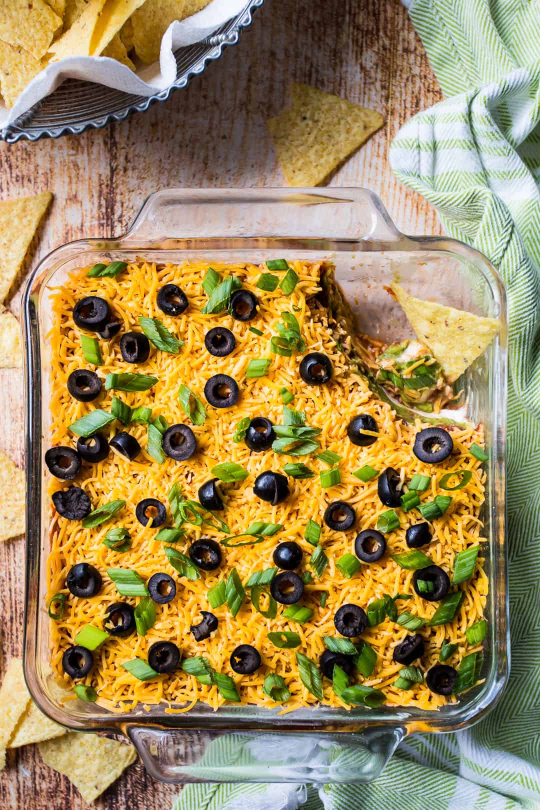 Overhead image of 7-layer dip recipe, with a basket of corn chips and a green kitchen towel.
