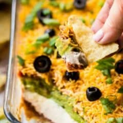 Close-up image of a tortilla chip taking a scoop out of a dish of 7-layer Mexican dip, with a text overlay above that reads "7-Layer Dip."