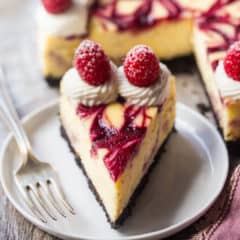 A perfect slice of white chocolate raspberry cheesecake on a white plate, with the whole cheesecake in the background.