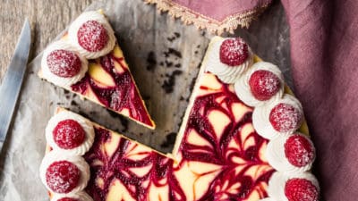 Overhead image of a white chocolate raspberry cheesecake sliced, with a soft pink and gold napkin.