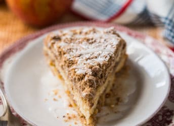 Slice of apple coffee cake on a red patterned plate, with fresh apples in the background.