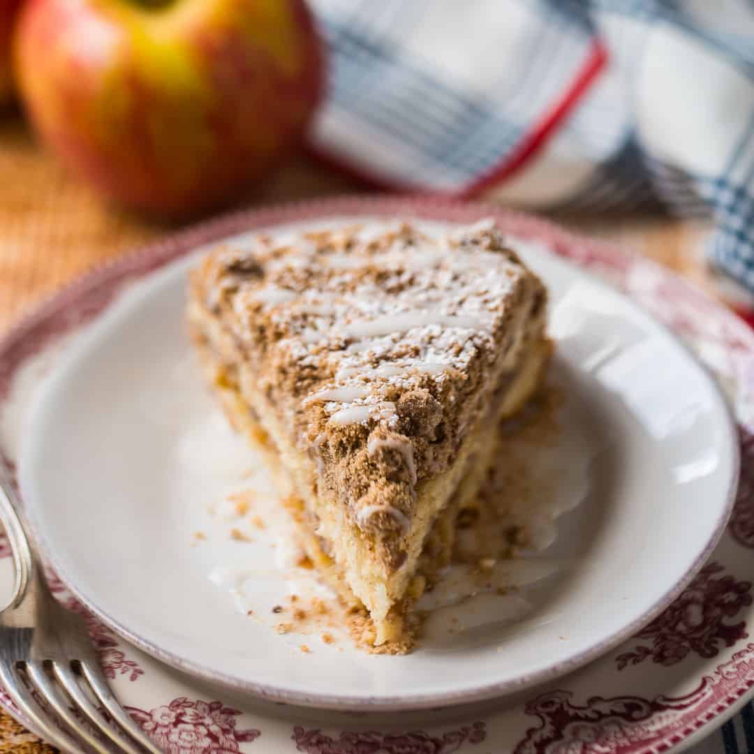 Slice of apple coffee cake on a red patterned plate, with fresh apples in the background.