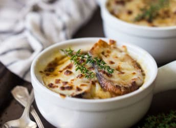 French onion soup in a white ceramic crock on a dark brown tray with vintage silver spoons.