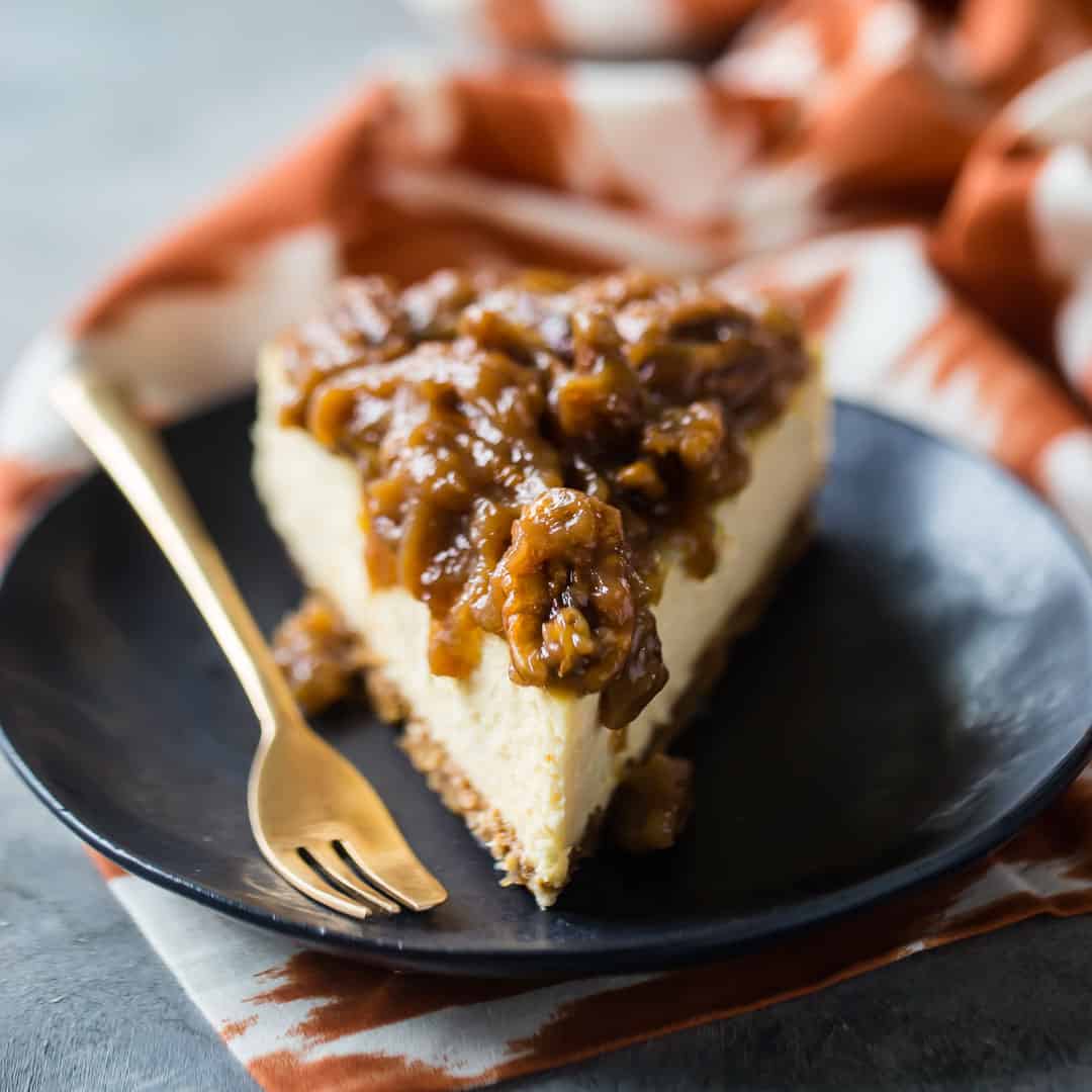 Slice of pecan pie cheesecake on a dark plate with a gold fork.