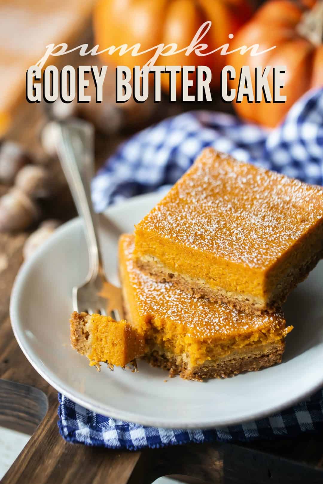 Pumpkin gooey cake bars on a white plate with a blue checked napkin and a text overlay above that reads "Pumpkin Gooey Butter Cake."
