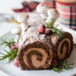Yule log cake garnished with greens & berries, on a white platter.