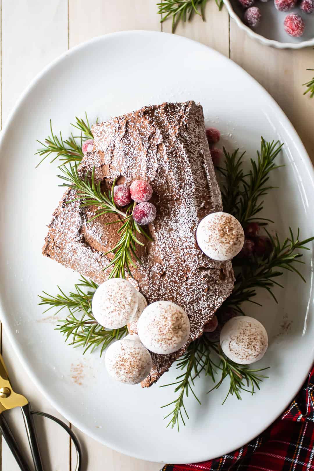 Overhead image of a chocolate yule log buche de noel cake garnished with powdered sugar, meringue mushrooms, greens, and sugared cranberries.