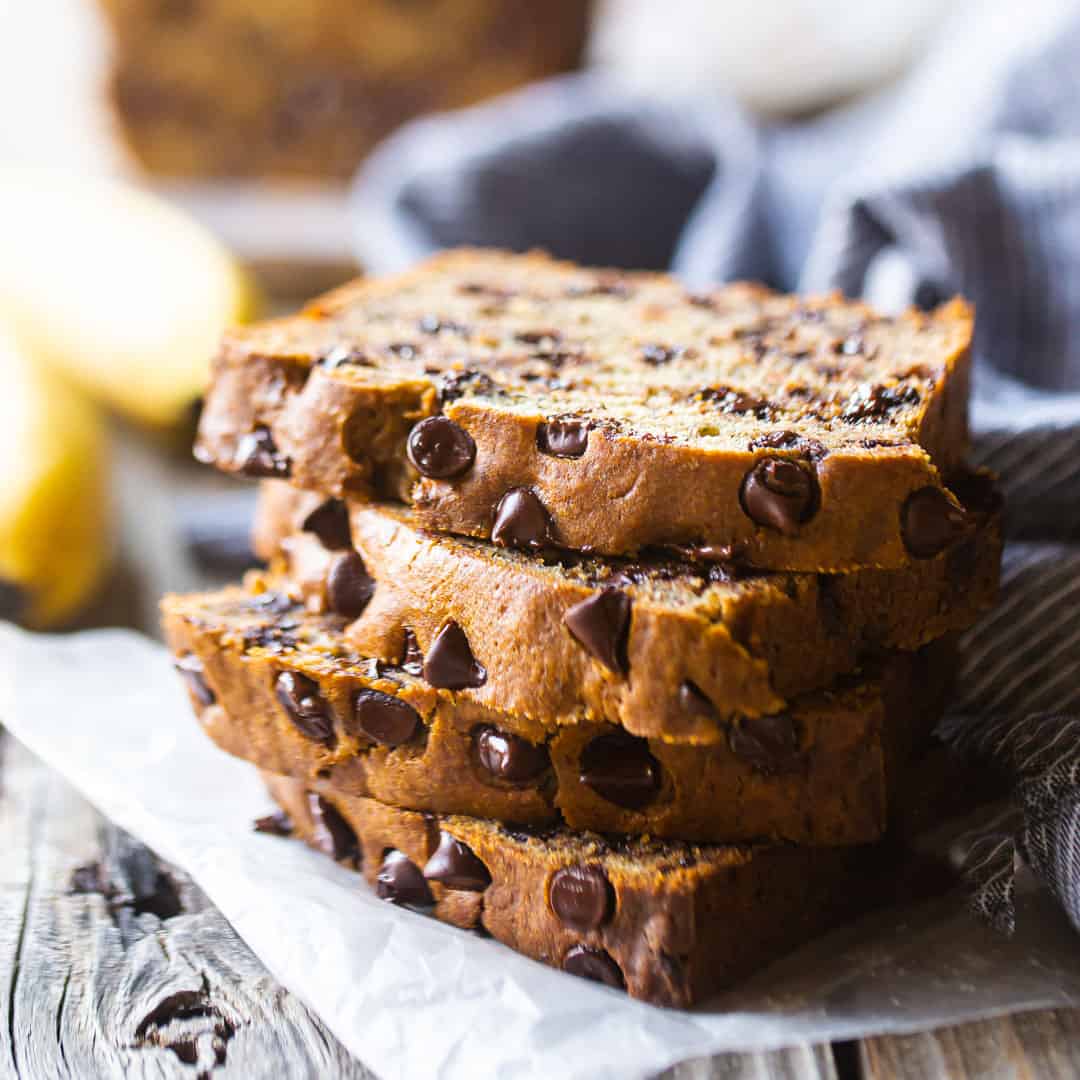 Chocolate chip banana bread slices stacked on a wood table with fresh bananas in the background.