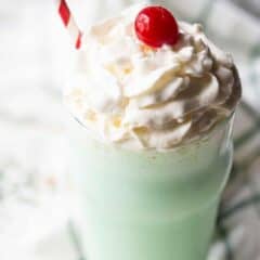 Shamrock shake recipe prepared and served in a glass with whipped cream, with a green plaid napkin in the background.