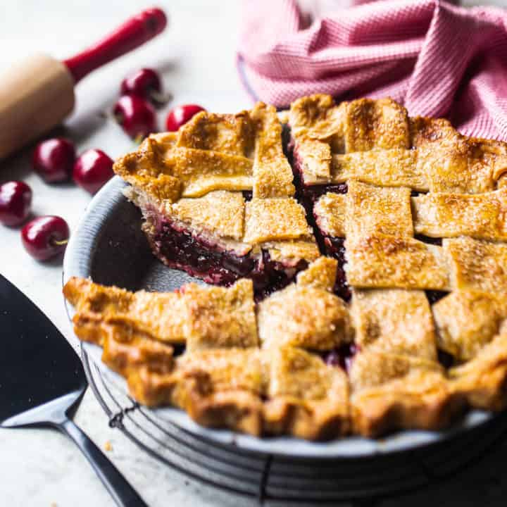 Cherry pie with a few slices cut, on a wire cooling rack with fresh cherries in the background.