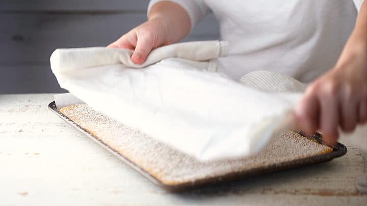 Laying a clean kitchen towel on top of a freshly baked pumpkin roll cake.