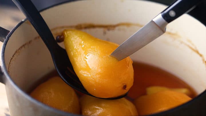 Sliding the tip of a sharp knife into a poached pear to check for doneness.