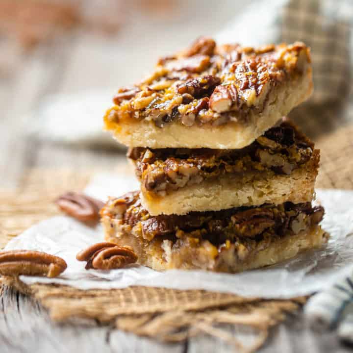 Pecan pie bars stacked on waxed paper, with whole pecans in the background.