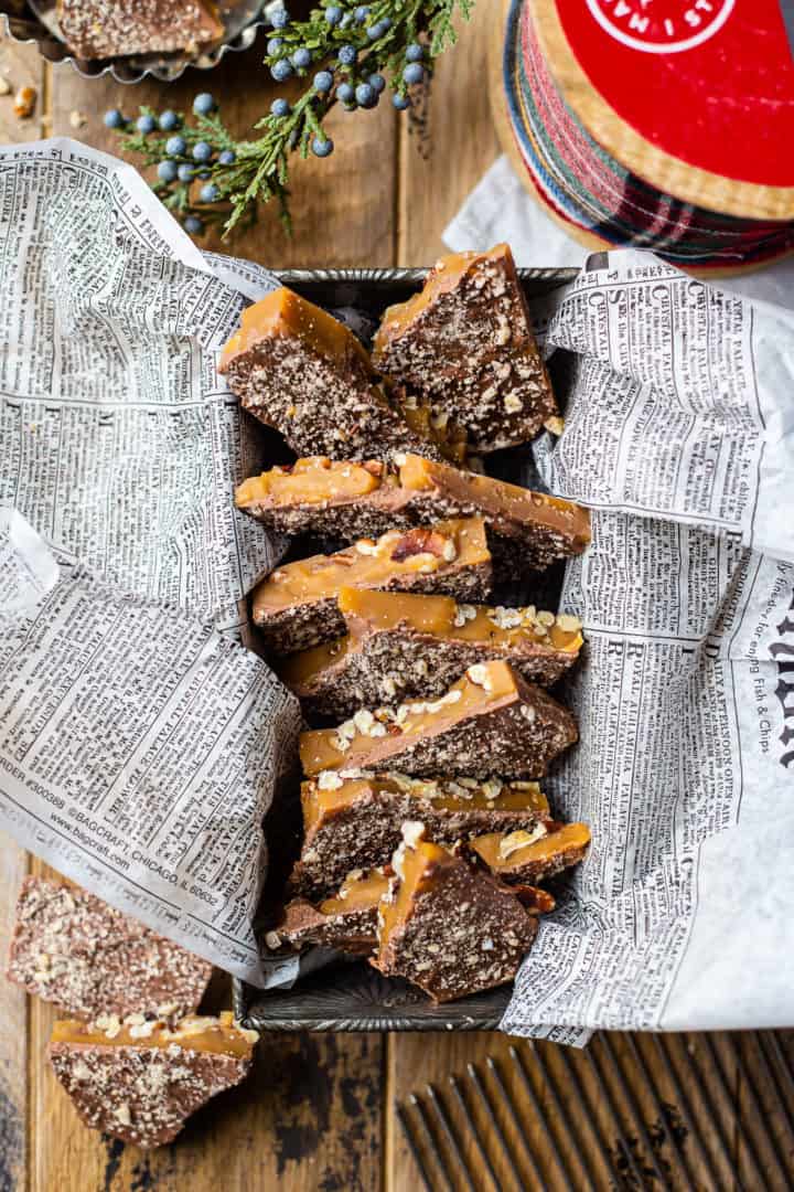 Overhead image of thick shards of homemade toffee, lined up in an antique loaf pan, surrounded with greenery and ribbon.