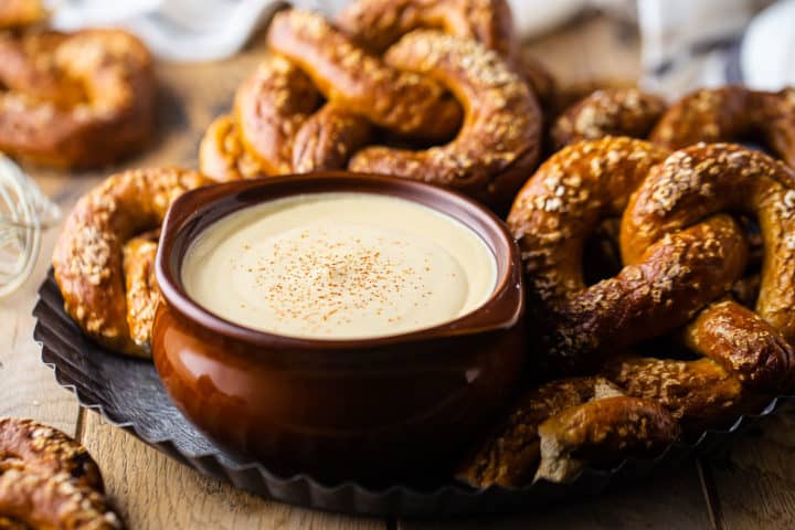 Beer cheese dip recipe, prepared and served in a pot-bellied brown bowl, with soft pretzels in the background.