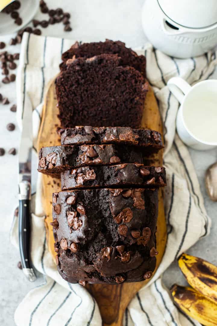 Overhead image of chocolate banana bread recipe, prepared and sliced, surrounded with bananas and chocolate chips.