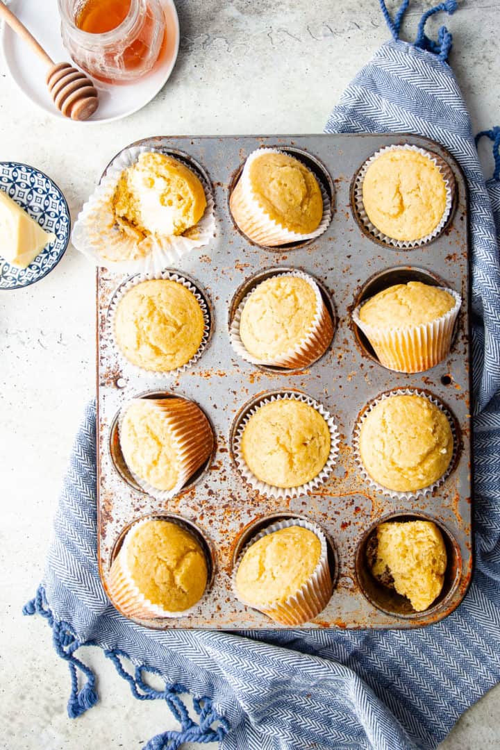 Overhead image of corn muffins arranged on a vintage pan, with a jar of honey and a blue kitchen cloth.