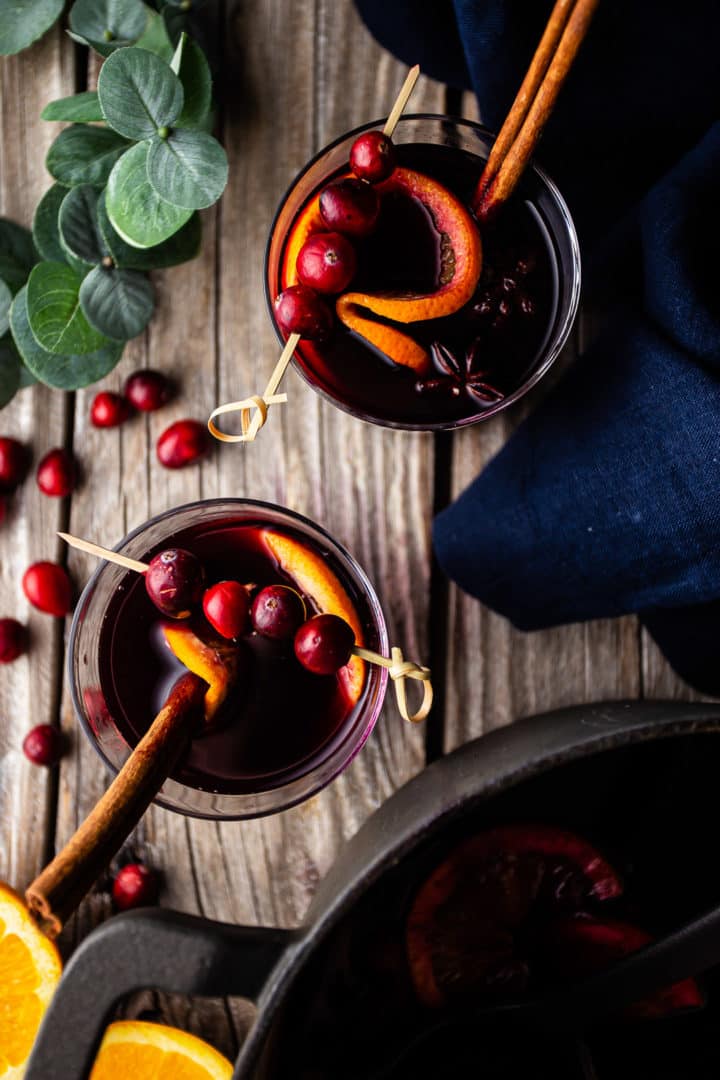 Overhead image of how to make mulled wine, served from a pot into two glasses and garnished with fresh cranberries and orange slices.