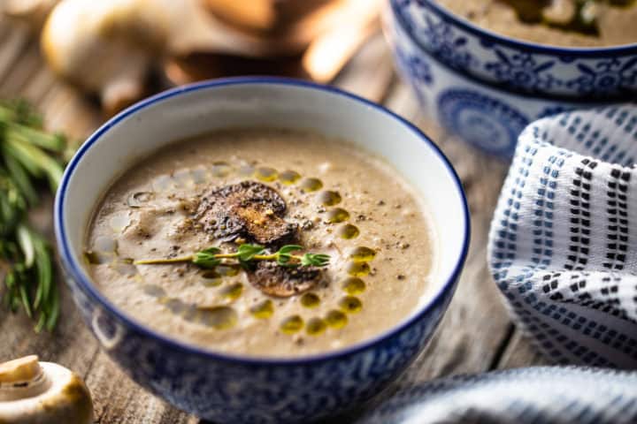 Close-up image of cream of mushroom soup recipe, prepared and garnished with cracked black pepper, truffle oil, and a sprig of thyme.