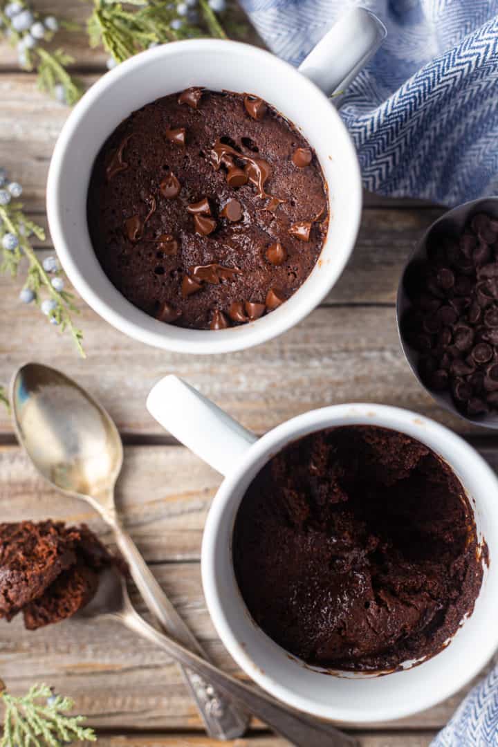 Overhead image of two chocolate cake in a mug served on a distressed wooden surface with vintage silver spoons.