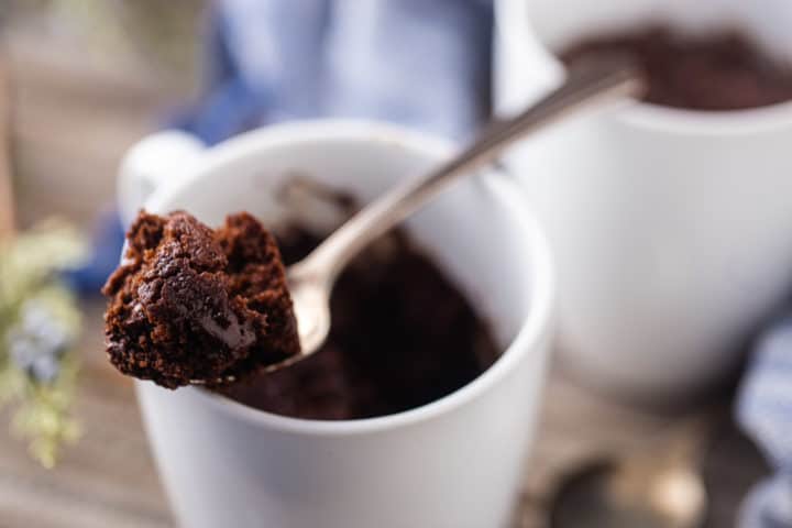Close-up image of a spoonful of mug cake chocolate resting on the rim of a white ceramic coffee cup.