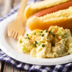 A scoop of potato salad on a paper plate with a hot dog in the background.