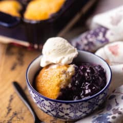 Square image of a bowl of blueberry cobbler topped with a scoop of vanilla ice cream.
