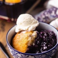 Square image of a bowl of blueberry cobbler topped with a scoop of vanilla ice cream.