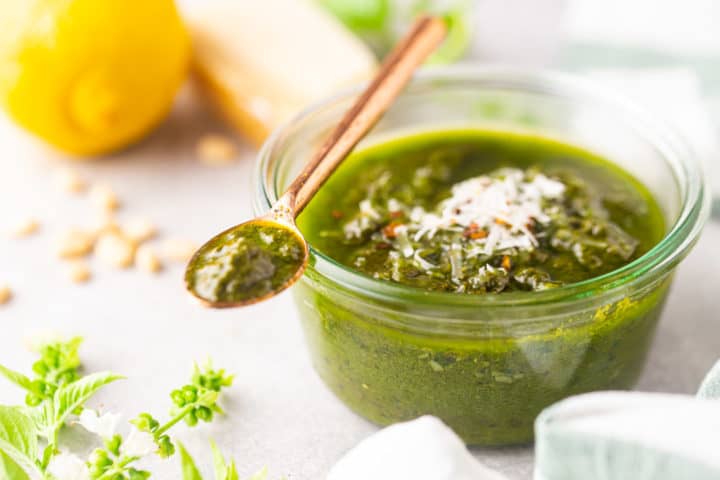 A jar of basil pesto on a pale background, surrounded by fresh basil, lemon, parmesan, and pine nuts.