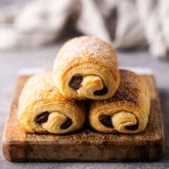 Three pain au chocolat stacked on a wooden serving board.