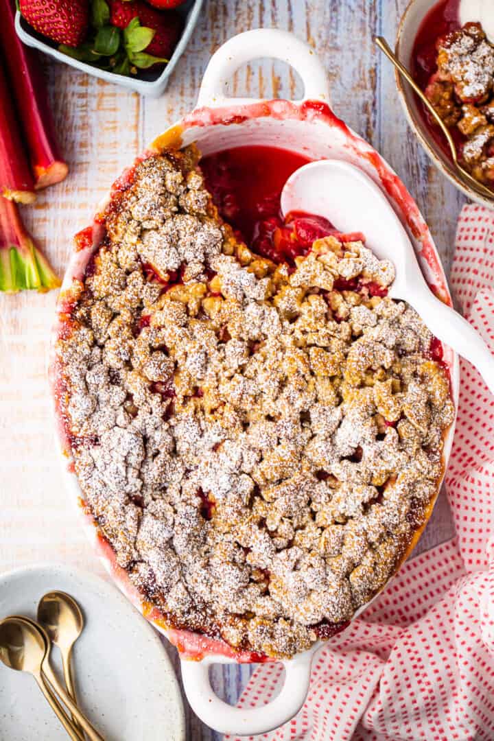Overhead image of strawberry rhubarb crisp recipe, prepared and baked in an oval dish and surrounded with fresh strawberries and rhubarb stalks.